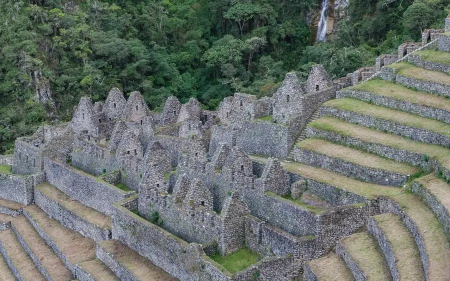 Rangées de ruines de pierre à pignon le long de terrasses herbeuses, une cascade dans la forêt en arrière-plan